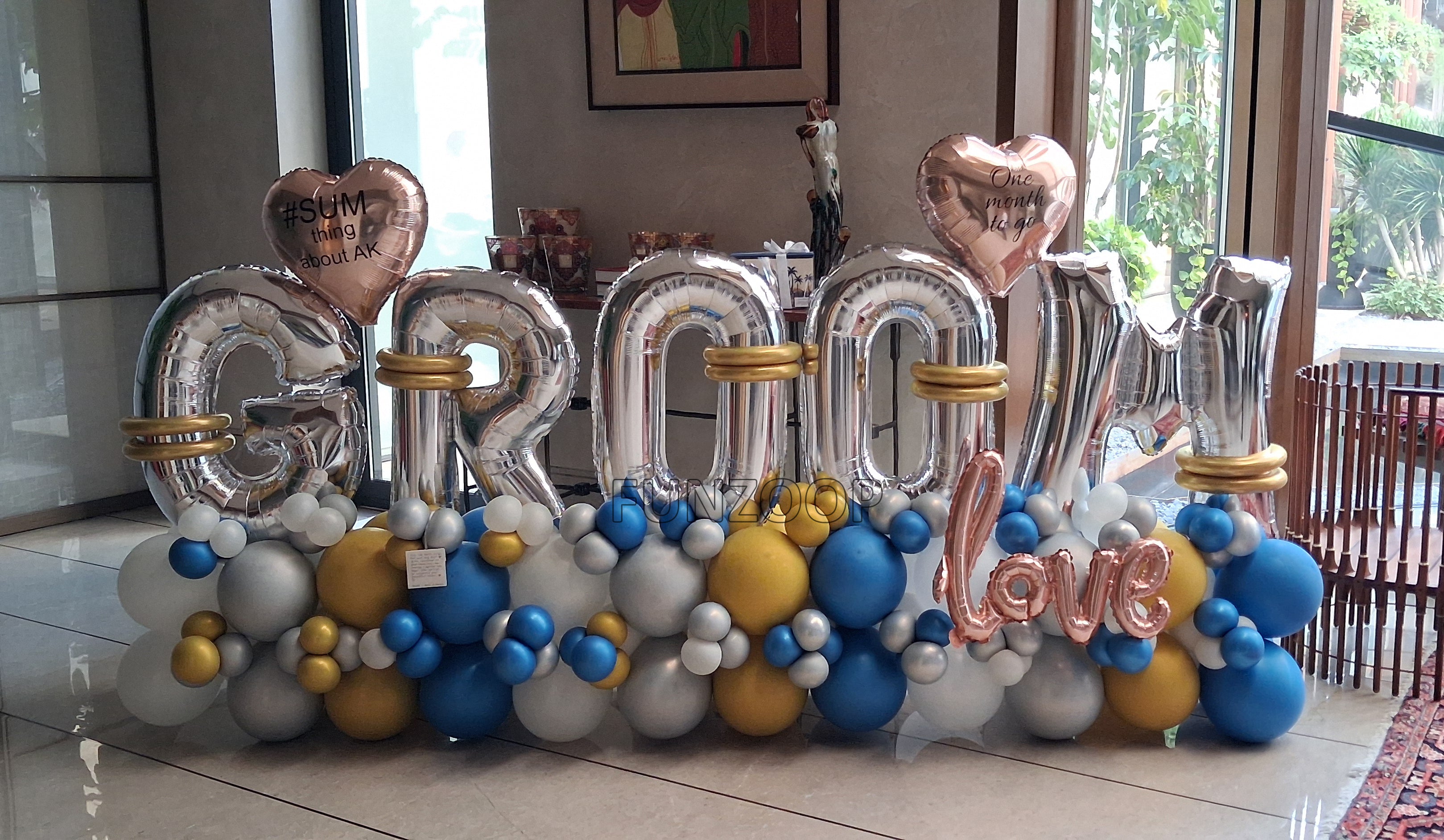 Silver GROOM balloon letters with personalised hearts, rose gold LOVE lettering and blue, gold, white and silver balloons placed indoor.