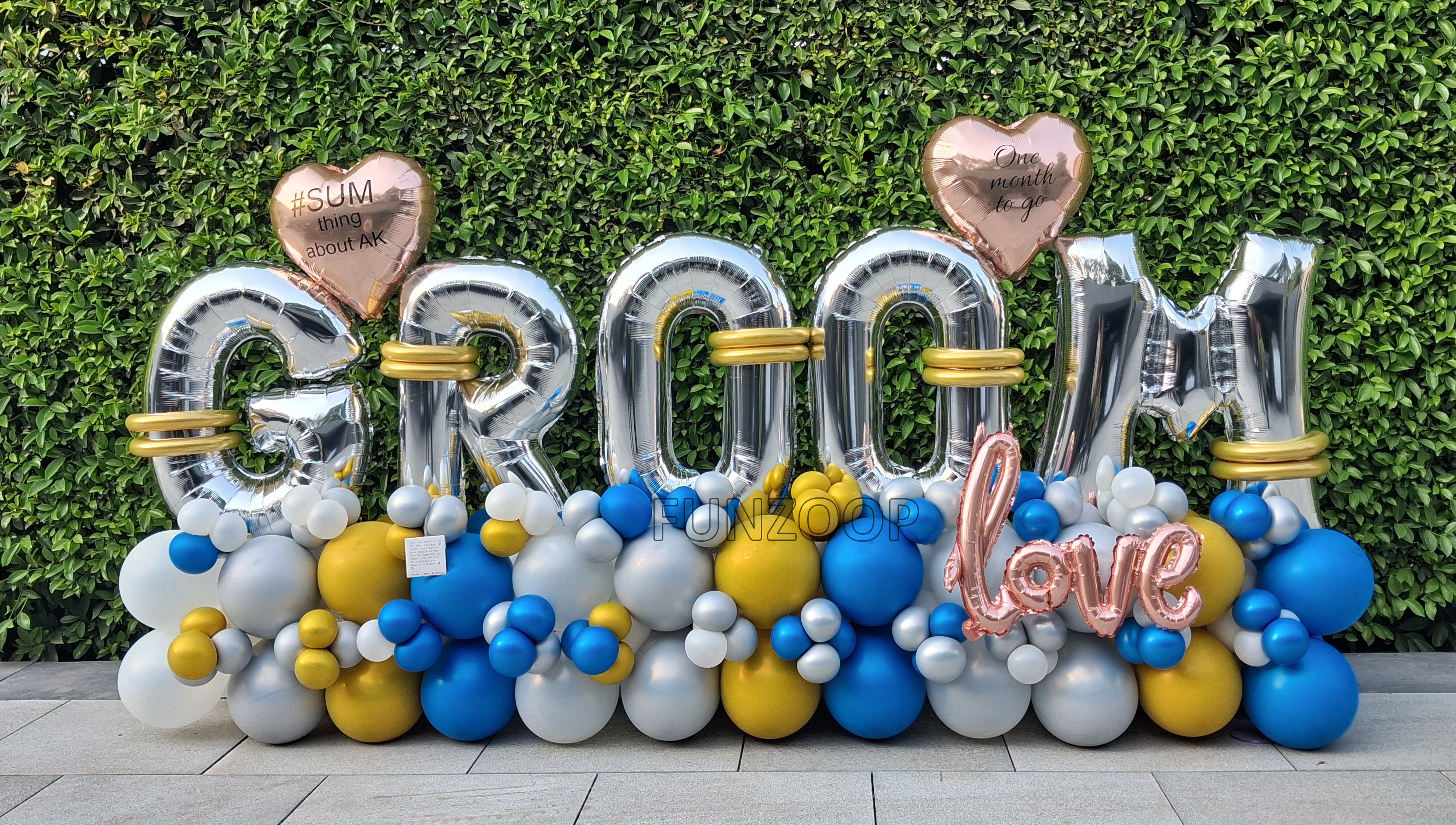 Silver GROOM balloon letters with personalised hearts, rose gold LOVE lettering and blue, gold, white and silver balloons placed outdoor.