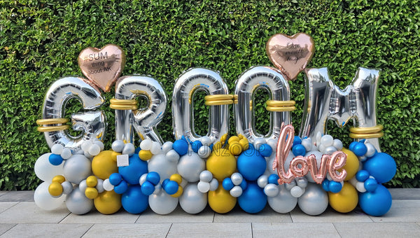 Silver GROOM balloon letters with personalised hearts, rose gold LOVE lettering and blue, gold, white and silver balloons placed outdoor.