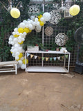 Asymmetric white and yellow balloon arch beside a white open-frame table with bunting in a leafy outdoor setting.