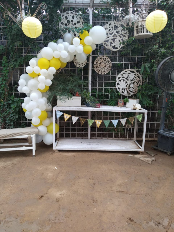 Asymmetric white and yellow balloon arch beside a white open-frame table with bunting in a leafy outdoor setting.